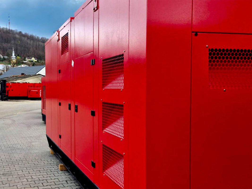 Side view of multiple red Zenessis generators lined up outdoors. The generators have visible ventilation grilles on the sides, and the surrounding area features a mountainous background with houses and a communication tower in the distance.