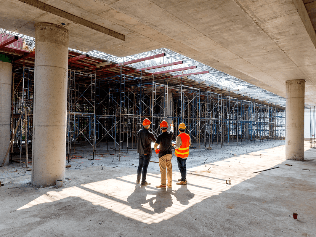 Trei muncitori în echipament de protecție (căști și veste reflectorizante) inspectează structura unui șantier de construcții aflat sub un planșeu de beton, cu schele metalice și piloni de susținere vizibili.