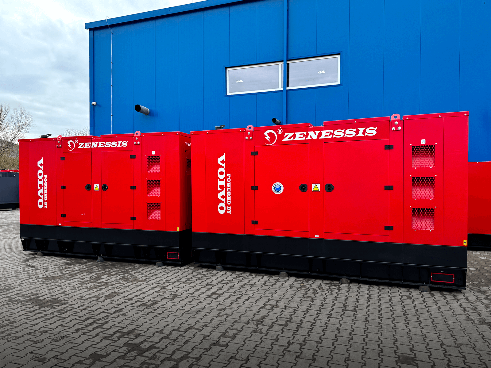 Two red Zenessis generators positioned side by side in front of a blue building. One generator has its door open, revealing the engine inside. Both units are branded with the Zenessis logo, and the 'Powered by Volvo' label is visible on the side of the second generator.